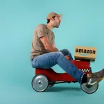 אדם בוגר נוסע על קטנוע צעצוע עם קופסא של אמזון למשלוח